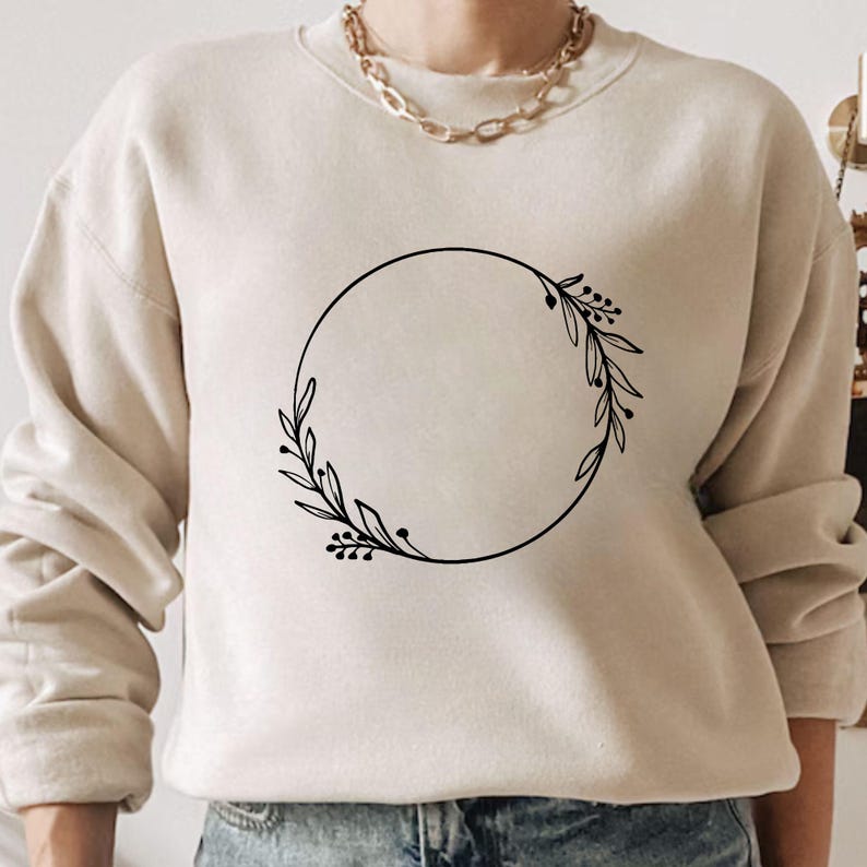 May include: Beige sweatshirt with a black circular floral wreath design. The wreath features leaves and berries. The sweatshirt has long sleeves and a crew neck. A gold chain necklace is visible at the neckline.