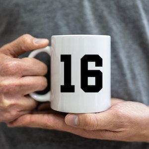 May include: White ceramic mug featuring the number 16 in a bold, black typeface. The mug is held in a person's hands, with a plain grey background. The mug is a standard size, ideal for hot beverages like tea or coffee.