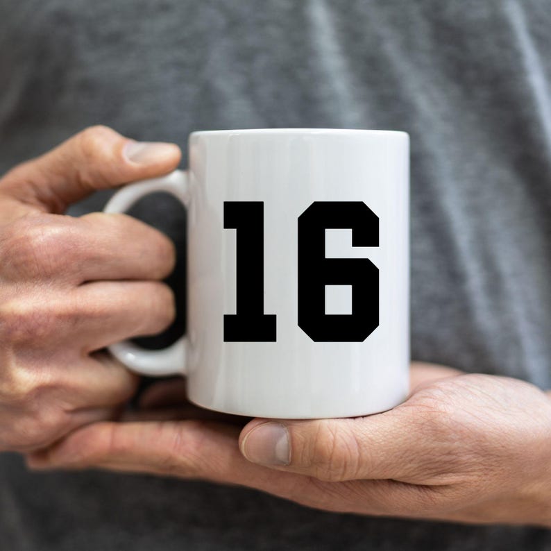 May include: White ceramic mug featuring the number 16 in a bold, black typeface. The mug is held in a person's hands, with a plain grey background. The mug is a standard size, ideal for hot beverages like tea or coffee.