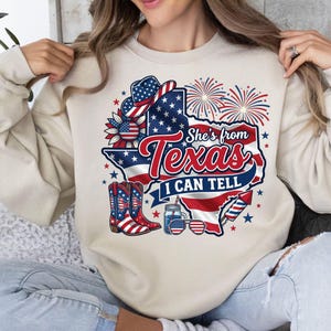 Pode incluir: Uma camisola bege de gola redonda com um gr&aacute;fico do estado do Texas preenchido com um padr&atilde;o de bandeira vermelha, branca e azul. O design inclui um chap&eacute;u de cowboy, girassol, botas, fogo de artif&iacute;cio, &oacute;culos e o texto She's from Texas I CAN TELL.