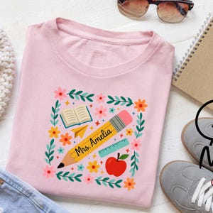 May include: A light pink t-shirt featuring a central graphic of a yellow pencil labeled "Mrs. Amelia," a red apple, an open book, a paper airplane, and a ruler, all framed by a colorful border of small flowers and green leafy vines.