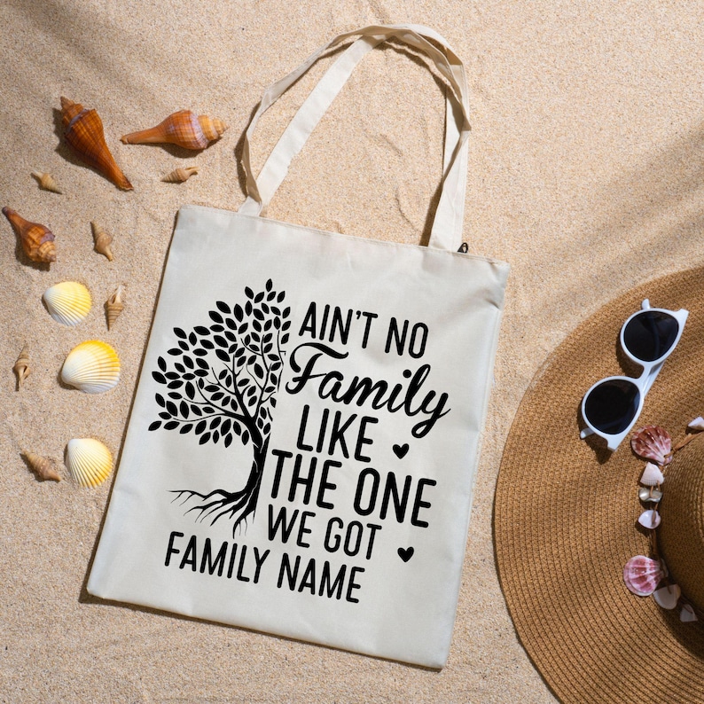 Pu&ograve; includere: Borsa tote color crema con la scritta "Ain't No Family Like The One We Got Family Name" e una grafica ad albero. La borsa ha manici lunghi ed &egrave; appoggiata sulla sabbia, accanto a un cappello di paglia e conchiglie. Gli occhiali da sole sono bianchi.