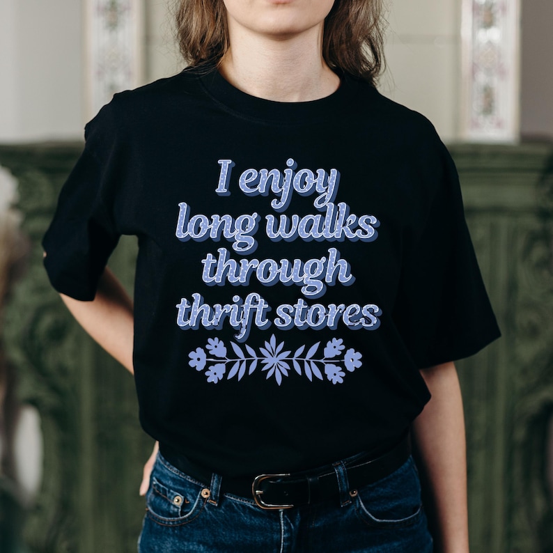 Może przedstawiać: Czarny t-shirt z napisem "I enjoy long walks through thrift stores" w jasnoniebieskiej, tr&oacute;jwymiarowej czcionce. Jasnoniebieski kwiatowy wz&oacute;r znajduje się na dole tekstu. Koszulka jest noszona przez osobę.
