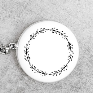 May include: A white, round keychain pendant featuring a black, minimalist wreath design. The wreath is composed of delicate leaves and small berries, encircling a blank space. A silver chain is attached to the pendant.