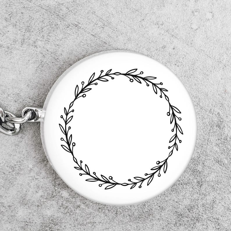 May include: A white, round keychain pendant featuring a black, minimalist wreath design. The wreath is composed of delicate leaves and small berries, encircling a blank space. A silver chain is attached to the pendant.