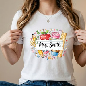 May include: A white short-sleeved t-shirt featuring a colorful graphic of school supplies including a red apple, a yellow pencil, a ruler, paper clips, and a coffee cup. A central white paper strip displays the black text Mrs. Smith.