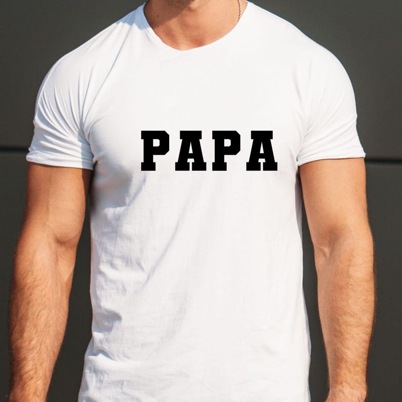 May include: White t-shirt featuring the word "PAPA" in large, black, block letters. The shirt has a crew neck and short sleeves. The image shows the upper body of a person wearing the shirt, with the text prominently displayed.