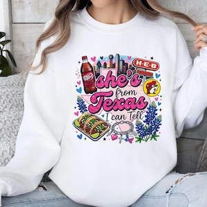 Pode incluir: Uma camisola branca de gola redonda com uma impress&atilde;o gr&aacute;fica colorida com o texto 'she's from Texas I can Tell'. O design inclui uma garrafa de Dr Pepper, um horizonte da cidade, um sinal H-E-B, uma mascote castor, tacos, tremo&ccedil;os e uma pulseira com berloques.
