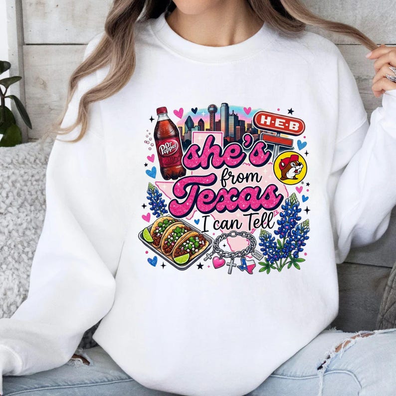 Pode incluir: Uma camisola branca de gola redonda com uma impress&atilde;o gr&aacute;fica colorida com o texto 'she's from Texas I can Tell'. O design inclui uma garrafa de Dr Pepper, um horizonte da cidade, um sinal H-E-B, uma mascote castor, tacos, tremo&ccedil;os e uma pulseira com berloques.