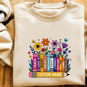 May include: A beige crewneck sweatshirt featuring a graphic of colorful books and flowers. The book spines are labeled with the words TEACH, LOVE, INSPIRE, LISTEN, MOTIVATE, LEAD, CONNECT, INCLUDE, HOME, CARE, and ENCOURAGE. A pencil graphic at the bottom reads CUSTOM NAME.