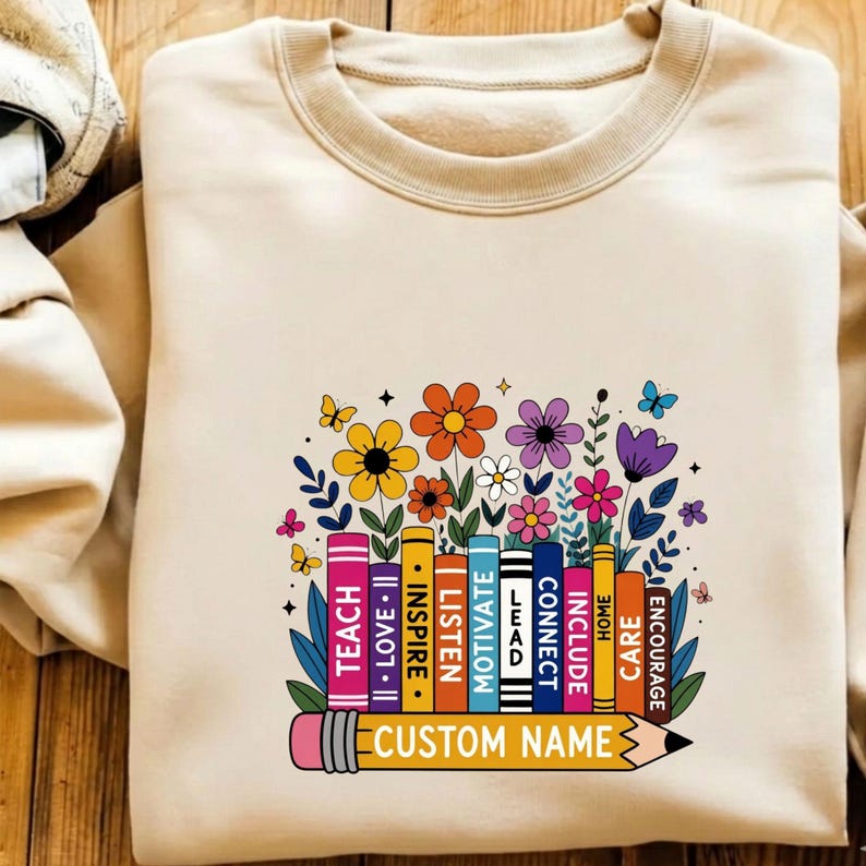 May include: A beige crewneck sweatshirt featuring a graphic of colorful books and flowers. The book spines are labeled with the words TEACH, LOVE, INSPIRE, LISTEN, MOTIVATE, LEAD, CONNECT, INCLUDE, HOME, CARE, and ENCOURAGE. A pencil graphic at the bottom reads CUSTOM NAME.