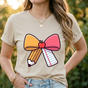 May include: A beige short-sleeve t-shirt featuring a graphic of a large bow. The left side of the bow is a yellow pencil, and the right side is a piece of lined notebook paper. A small red apple is centered at the knot of the bow.