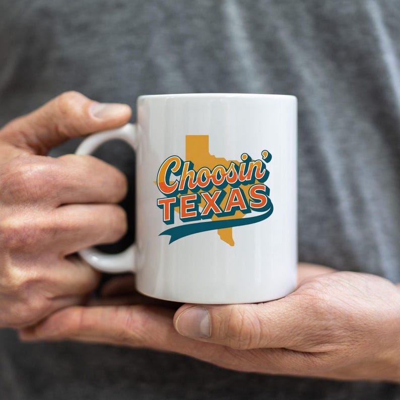 May include: White ceramic mug with the text "Choosin' TEXAS" in orange, teal, and yellow. The design includes a yellow outline of the state of Texas. The mug is held by a person.