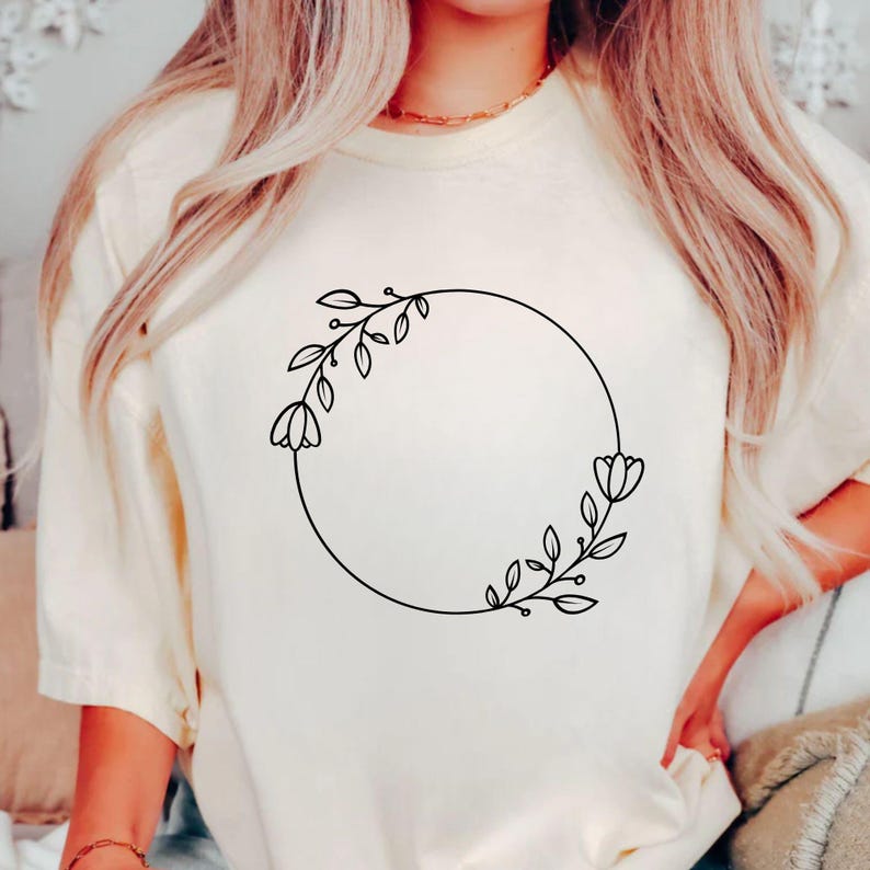 May include: Cream-colored t-shirt featuring a black floral wreath design. The wreath is a circular arrangement of leaves and flowers, with a minimalist, line-art style. The shirt has a relaxed fit and a soft, neutral color.