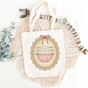 Może przedstawiać: Kremowa torba typu tote z ilustracją w stylu vintage przedstawiającą trzy r&oacute;żowe i białe miski do mieszania z kwiatowym wzorem. Projekt jest obramowany ozdobnym owalem z koronkową obw&oacute;dką i r&oacute;żową kokardą. Torba ma kremowe uchwyty.