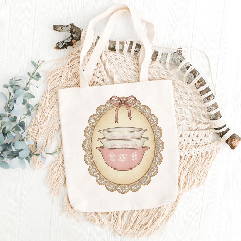 Może przedstawiać: Kremowa torba typu tote z ilustracją w stylu vintage przedstawiającą trzy r&oacute;żowe i białe miski do mieszania z kwiatowym wzorem. Projekt jest obramowany ozdobnym owalem z koronkową obw&oacute;dką i r&oacute;żową kokardą. Torba ma kremowe uchwyty.