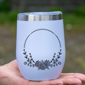 May include: A white insulated wine tumbler with a silver rim, held in a hand. The tumbler features a black floral design with a circular frame. The design includes flowers and leaves, adding a decorative touch to the drinkware.