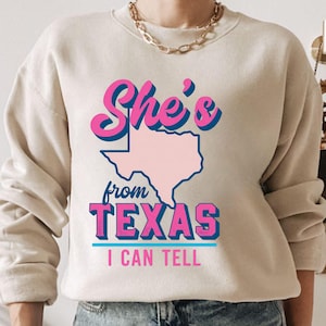 May include: Beige sweatshirt with the text "She's from TEXAS I CAN TELL" in pink and blue lettering. The word "TEXAS" is below a pink outline of the state. The sweatshirt has long sleeves and a crew neck.