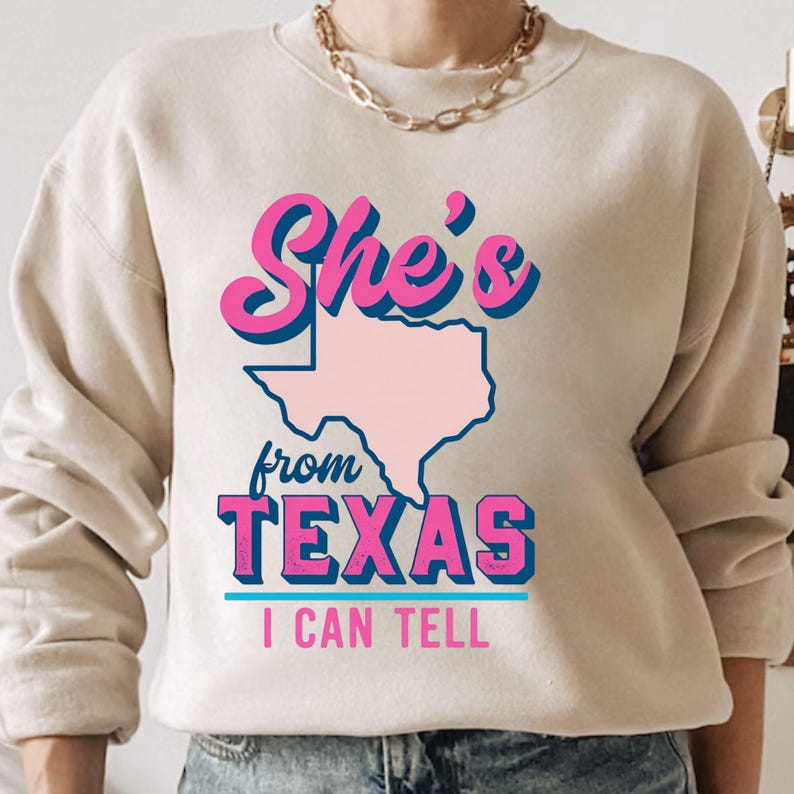 May include: Beige sweatshirt with the text "She's from TEXAS I CAN TELL" in pink and blue lettering. The word "TEXAS" is below a pink outline of the state. The sweatshirt has long sleeves and a crew neck.
