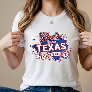 Pode incluir: Uma t-shirt branca de manga curta com um gr&aacute;fico do estado do Texas em glitter azul. O texto diz She's from TEXAS I CAN TELL com detalhes em vermelho e estrelas. Um log&oacute;tipo circular da equipa de basebol Texas Rangers &eacute; vis&iacute;vel &agrave; direita.
