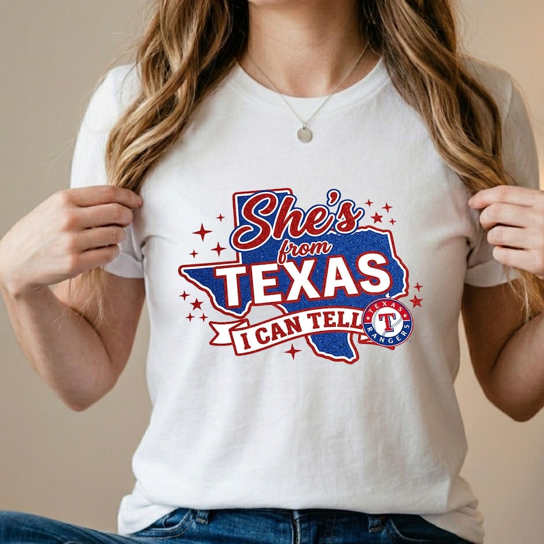 Pode incluir: Uma t-shirt branca de manga curta com um gr&aacute;fico do estado do Texas em glitter azul. O texto diz She's from TEXAS I CAN TELL com detalhes em vermelho e estrelas. Um log&oacute;tipo circular da equipa de basebol Texas Rangers &eacute; vis&iacute;vel &agrave; direita.
