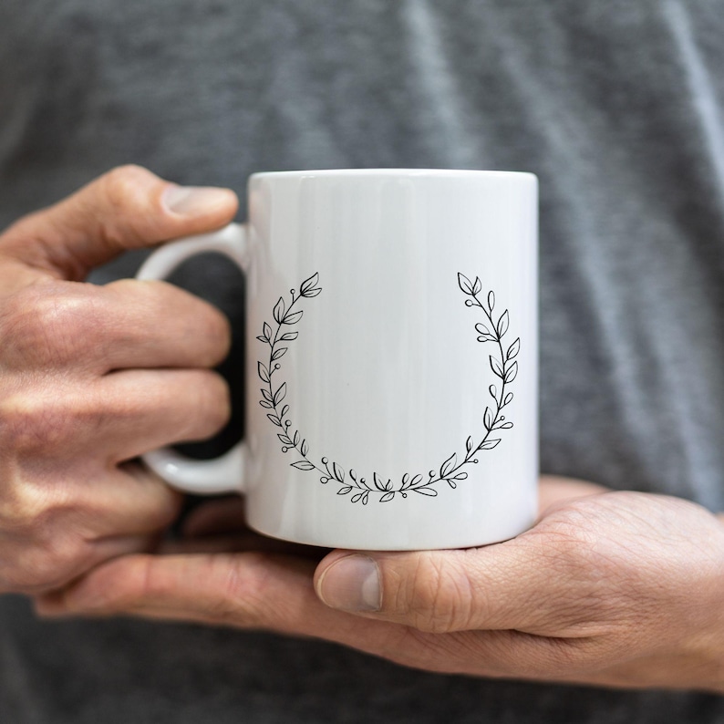 May include: White ceramic mug with a black floral wreath design. The mug has a curved handle and a cylindrical shape. The wreath is composed of leaves and small dots, creating a delicate, natural aesthetic. The mug is held in a person's hands.