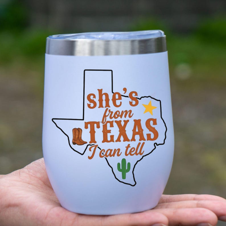 May include: White insulated wine tumbler with a silver rim. The tumbler features a Texas-shaped graphic with a cowboy boot, star, and cactus, along with the text "She's from Texas I can tell."