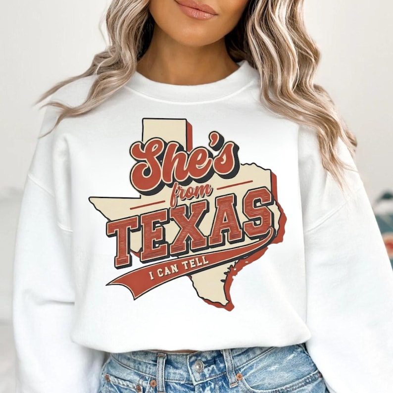 May include: White sweatshirt with the text "She's from Texas I can tell" in a retro design. The design features the outline of the state of Texas in a cream color with red and brown text.