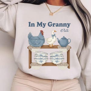 Może przedstawiać: Jasna bluza z napisem "In My Granny era". Grafika przedstawia niebieską kurę, białą gęś z niebieską kokardą, niebieski czajnik i kwiatowe miski na drewnianej p&oacute;łce, w stylu vintage.