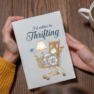 Może przedstawiać: Jasnoszara karta z napisem "I'd rather be Thrifting" w eleganckiej czcionce. Karta przedstawia w&oacute;zek sklepowy wypełniony przedmiotami w stylu vintage, takimi jak lampa, lustro i niebiesko-białe talerze, sugerując motyw zakup&oacute;w antyk&oacute;w.
