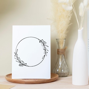 May include: A white card with a black floral wreath design, displayed on a wooden tray. The wreath is a simple circle with leaves and berries. Decorative vases with dried flowers are in the background.