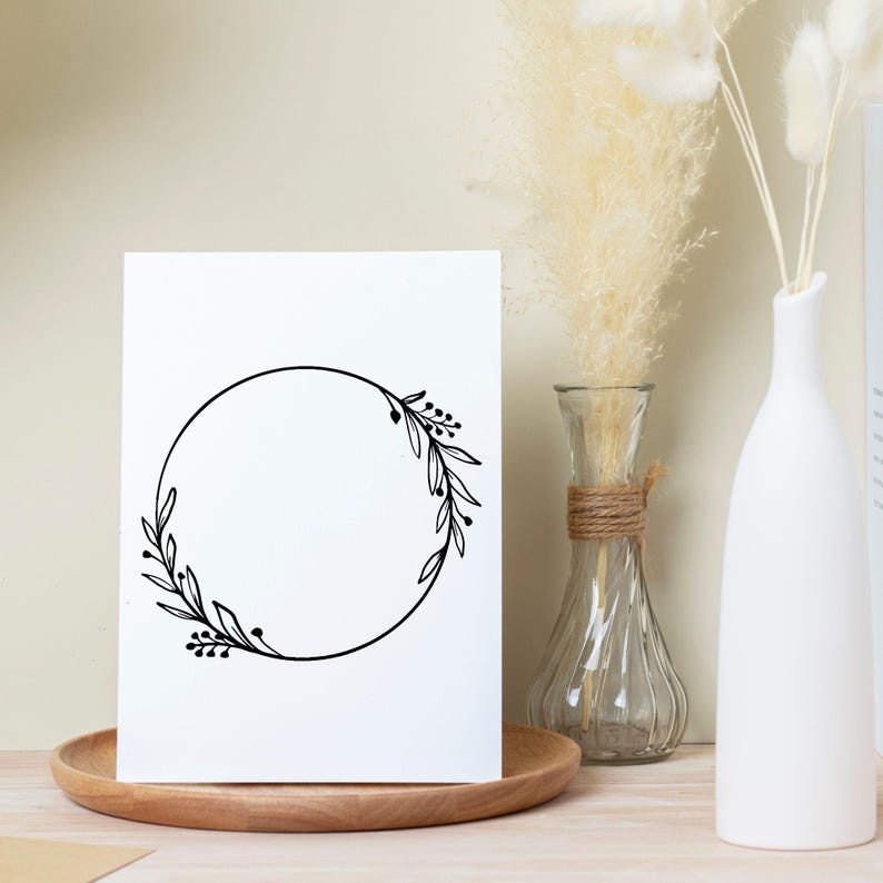 May include: A white card with a black floral wreath design, displayed on a wooden tray. The wreath is a simple circle with leaves and berries. Decorative vases with dried flowers are in the background.
