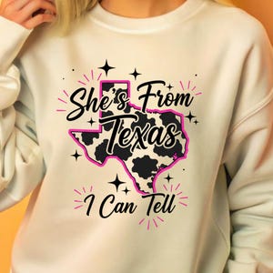 May include: Cream-colored sweatshirt with a graphic design featuring the state of Texas in pink, black, and white. The text "She's From Texas I Can Tell" is printed above and below the Texas outline, with star accents.