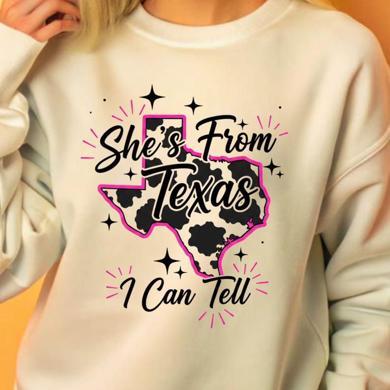 May include: Cream-colored sweatshirt with a graphic design featuring the state of Texas in pink, black, and white. The text "She's From Texas I Can Tell" is printed above and below the Texas outline, with star accents.