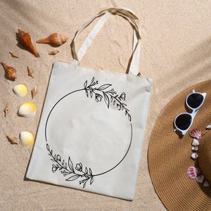 May include: A beige tote bag with a black floral wreath design. The bag has two handles and is lying on sand, next to seashells and a straw hat with sunglasses. The wreath is a circle of leaves and flowers.