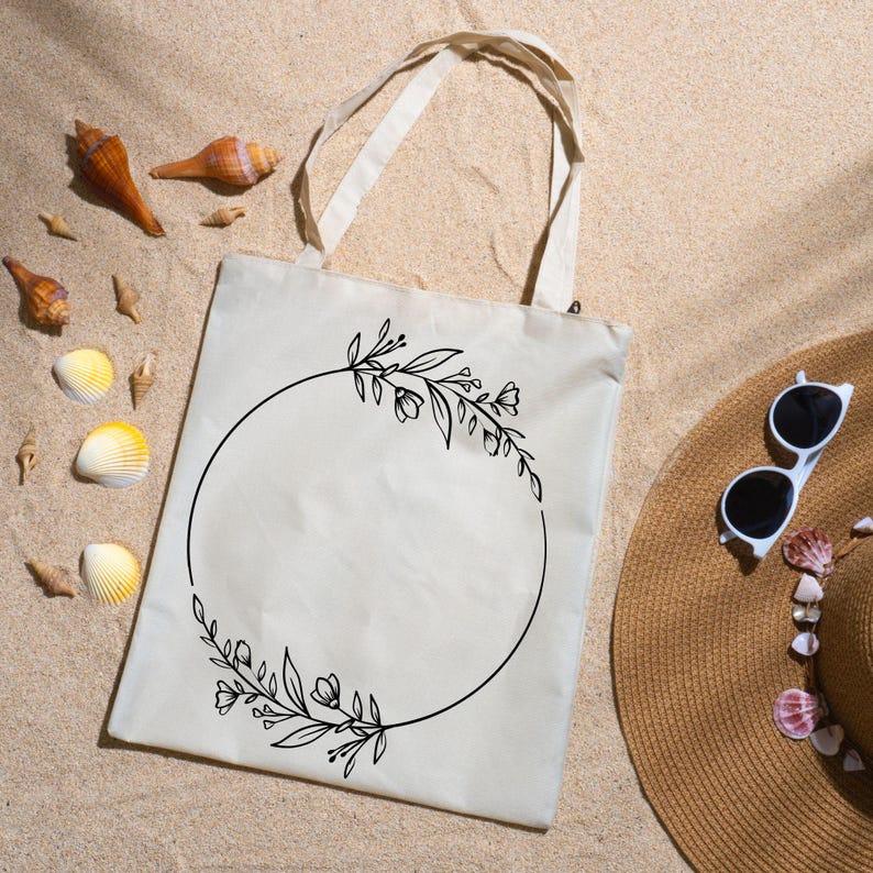 May include: A beige tote bag with a black floral wreath design. The bag has two handles and is lying on sand, next to seashells and a straw hat with sunglasses. The wreath is a circle of leaves and flowers.