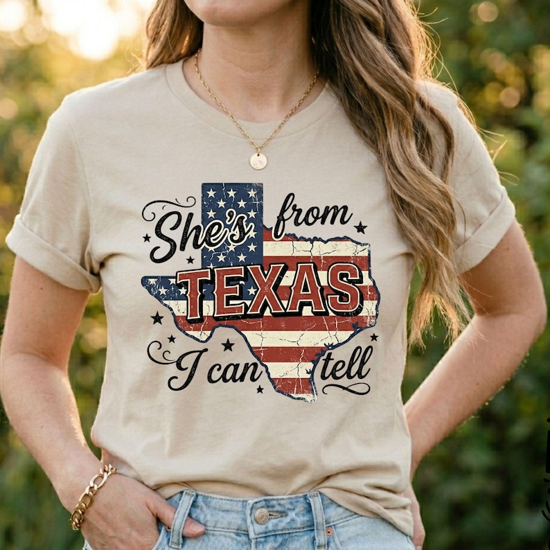 Pode incluir: Uma t-shirt bege de manga curta com um gr&aacute;fico do estado do Texas preenchido com o padr&atilde;o da bandeira americana em vermelho, branco e azul. O texto She's from TEXAS I can tell est&aacute; em preto com uma fonte desgastada e estilizada.