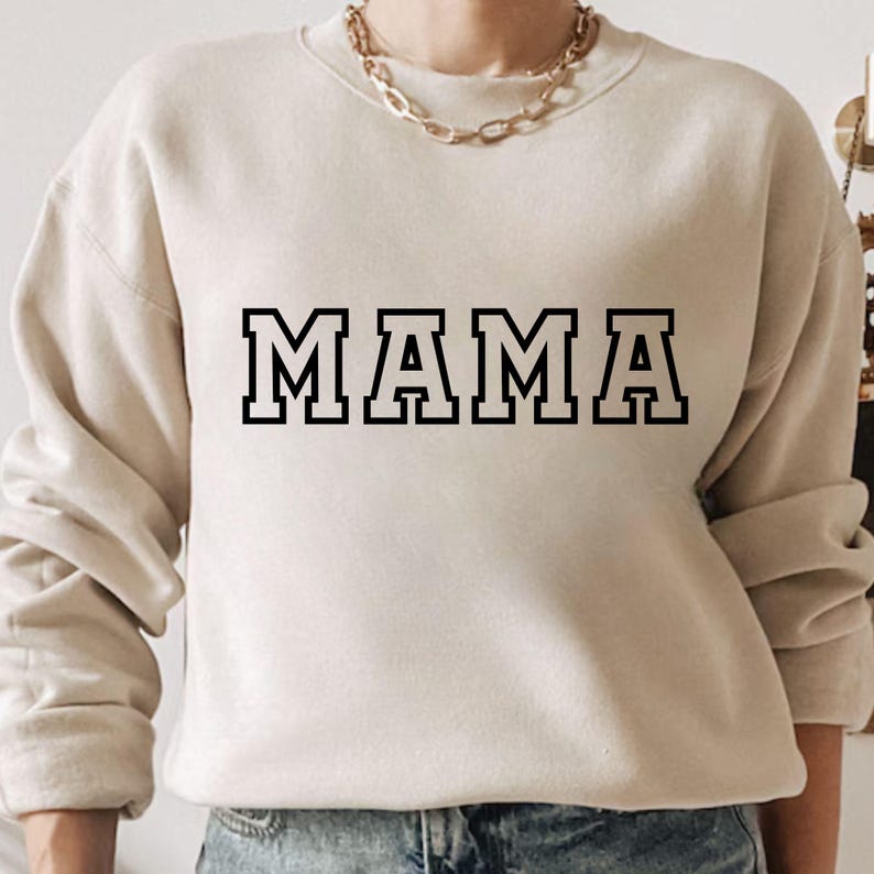 May include: Beige sweatshirt featuring the word "MAMA" in bold, black, block letters. The sweatshirt has long sleeves and a crew neck. A gold chain necklace is visible at the neckline. The sweatshirt is made of a soft material.