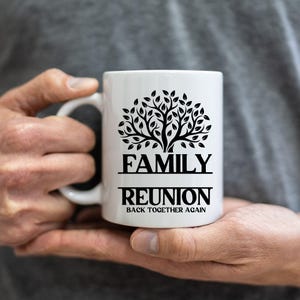 Pu&ograve; includere: Tazza in ceramica bianca con un disegno di albero nero e la scritta "FAMILY REUNION BACK TOGETHER AGAIN". La tazza ha un manico curvo ed &egrave; tenuta tra le mani di una persona.