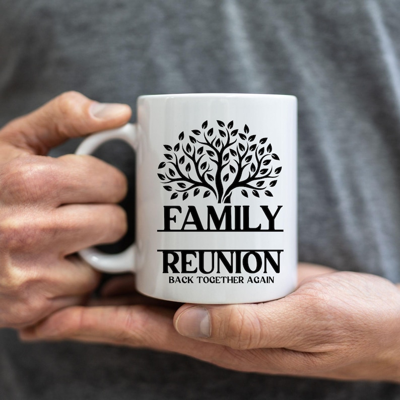 Pu&ograve; includere: Tazza in ceramica bianca con un disegno di albero nero e la scritta "FAMILY REUNION BACK TOGETHER AGAIN". La tazza ha un manico curvo ed &egrave; tenuta tra le mani di una persona.