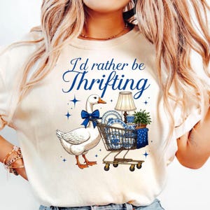 Może przedstawiać: Kremowa koszulka z niebieskim napisem "I'd rather be Thrifting". Kresk&oacute;wkowa gęś z niebieską kokardą ciągnie w&oacute;zek sklepowy wypełniony niebiesko-białą porcelaną, lampą i rośliną.