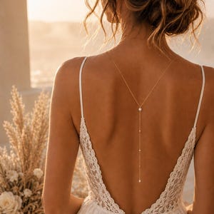 Peut inclure: Vue arrière d'une mariée en robe de mariée en dentelle blanche à dos nu. Un collier en or délicat avec une perle et de petites perles pend dans le dos. Les cheveux de la mariée sont coiffés en chignon avec une coiffe florale.