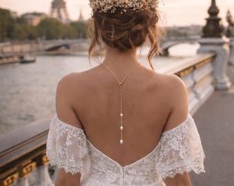 May include: A bride in a white lace off-the-shoulder wedding dress with a pearl back necklace and a floral crown. The Eiffel Tower is visible in the background, with a bridge and water.