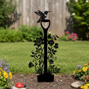 May include: Black metal garden stake featuring a hummingbird perched atop a shovel-shaped design. The stake is adorned with floral and vine details. The garden art is placed in a garden bed with flowers and grass.