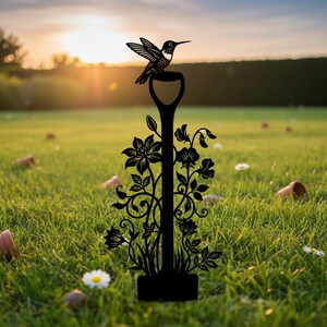 May include: A black metal garden stake featuring a hummingbird perched atop a shovel-shaped design. The stake is adorned with floral and vine details. The background shows a grassy lawn with a sunset.