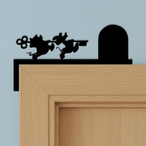 Peut inclure: Un ornement de porte en métal noir représentant la silhouette de deux souris transportant une grande clé vers une porte arrondie. Cette pièce décorative est conçue pour se poser sur le coin supérieur d'un cadre de porte.
