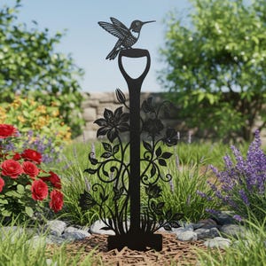 May include: Black metal garden stake featuring a hummingbird perched atop a shovel-shaped handle. The stake is adorned with floral and leaf cutouts. The garden art is placed in a garden setting with flowers and greenery.
