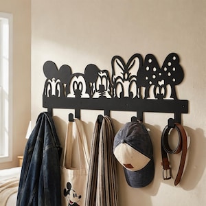 May include: A black metal wall-mounted coat rack featuring a decorative cutout design of five cartoon character heads along the top edge. The rack includes five downward-facing hooks currently holding a denim jacket, a tote bag, a scarf, a cap, and a belt.