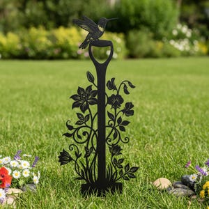 May include: Black metal garden stake featuring a hummingbird perched atop a shovel handle design. The stake is adorned with floral and leaf cutouts, adding a decorative touch to any outdoor space. The hummingbird has detailed wing and body features.