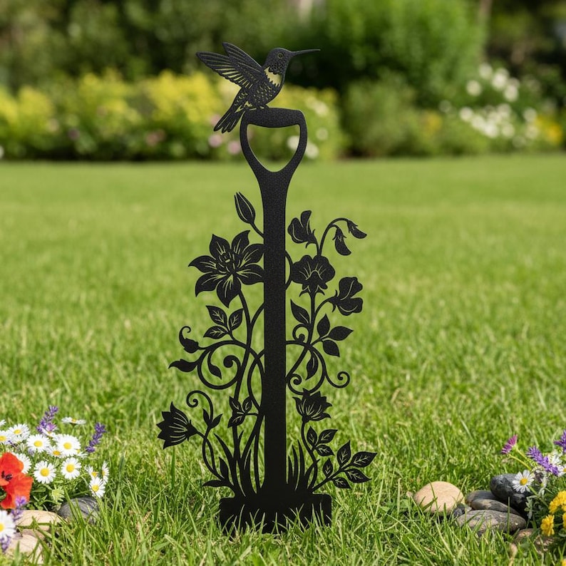 May include: Black metal garden stake featuring a hummingbird perched atop a shovel handle design. The stake is adorned with floral and leaf cutouts, adding a decorative touch to any outdoor space. The hummingbird has detailed wing and body features.